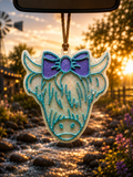 Highland Belle teal highland cow car freshie with purple bow hanging from rearview mirror at golden sunset