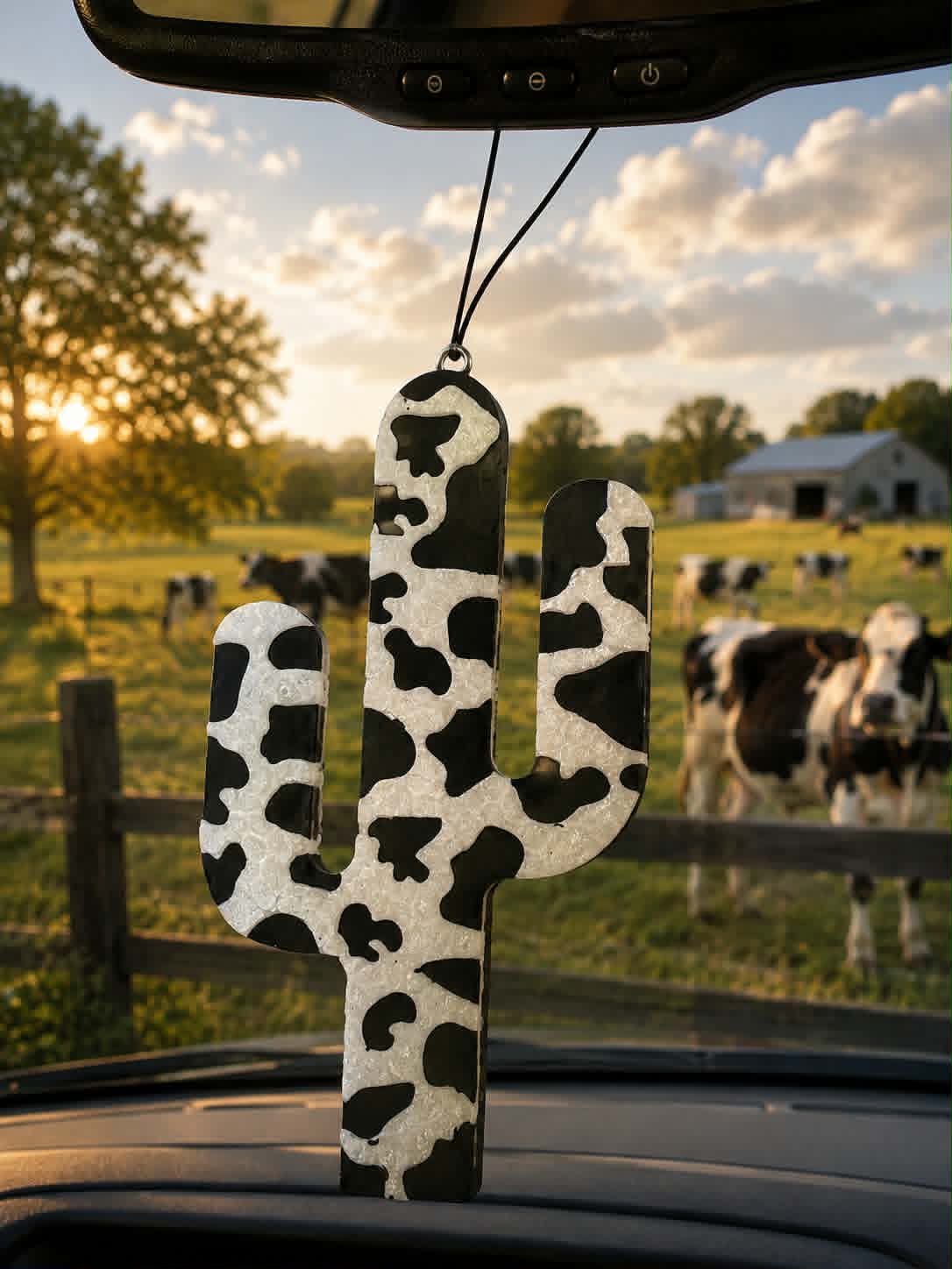 Cow print cactus car freshie hanging from rearview mirror with cattle in background